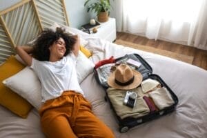 Happy woman lying down on bed next to open suitcase full of clothes, ready to go on vacation trip.