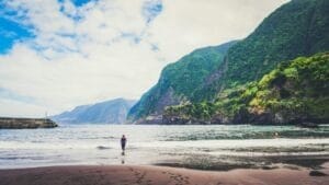Young girl in wetsuit goes into the sea water beach. It is alone surrounded by mountains of Madeira.