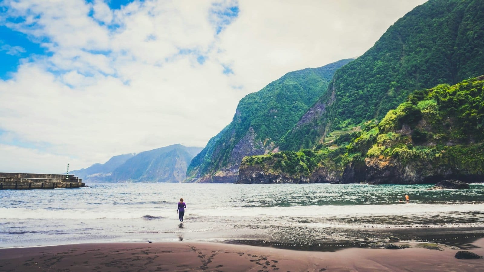 Young girl in wetsuit goes into the sea water beach. It is alone surrounded by mountains of Madeira.